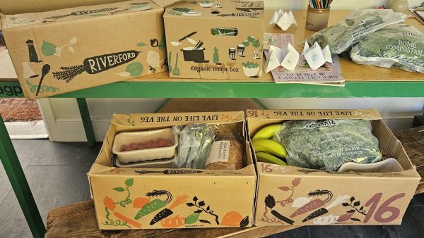 Riverford recipe and veg boxes unpacked on a table and bench, with kale, bananas, raspberries, bread and home-compostable bags.